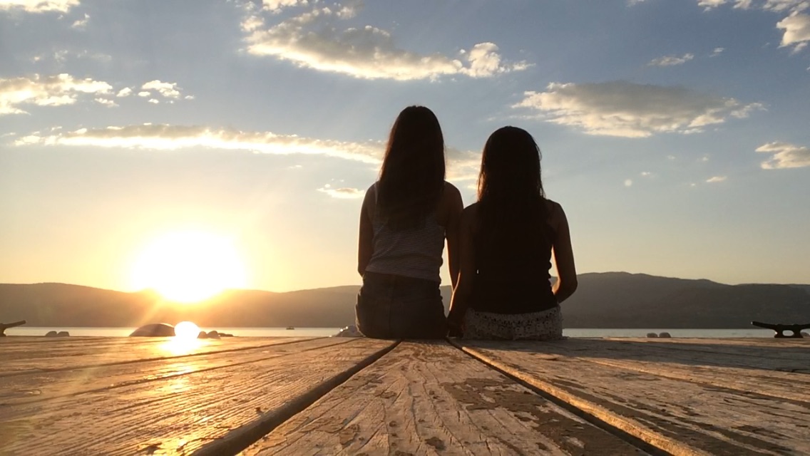 Two girls sitting on a wooden dock, watching the sun set over a lake